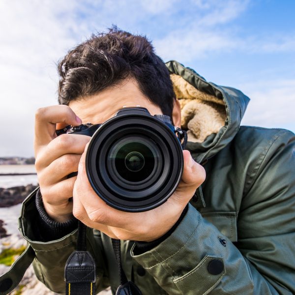 Eine Person mit kurzen braunen Haaren in einer dunkelgrünen Outdoorjacke hält eine Kamera vor sein Gesicht und fotografiert frontal.